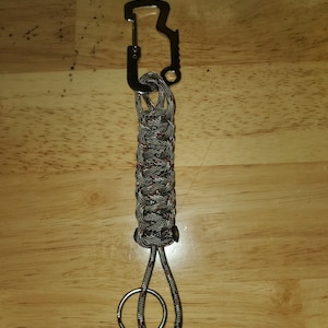 Puede incluir: Un llavero plateado con un abridor de botellas y un lazo de paracord trenzado. El lazo es de camuflaje con patrones marrones y grises.