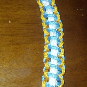 May include: A close-up of a braided cord with alternating blue, white, and gold sections. The cord is woven in a complex pattern, creating a decorative and durable design.