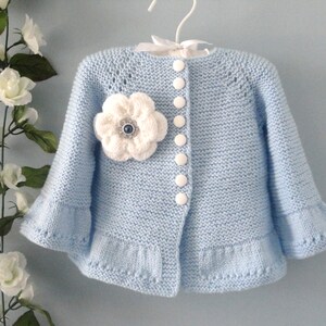 May include: A light blue knitted cardigan with a white flower embellishment and white buttons. The cardigan has a ruffled hem and sleeves.