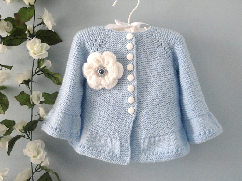 May include: A light blue knitted cardigan with a white flower embellishment and white buttons. The cardigan has a ruffled hem and sleeves.