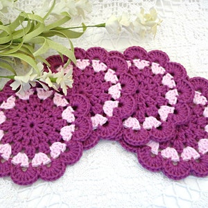 May include: Set of five crocheted coasters in shades of pink and purple. The coasters have a scalloped edge and a center design of pink and purple circles.