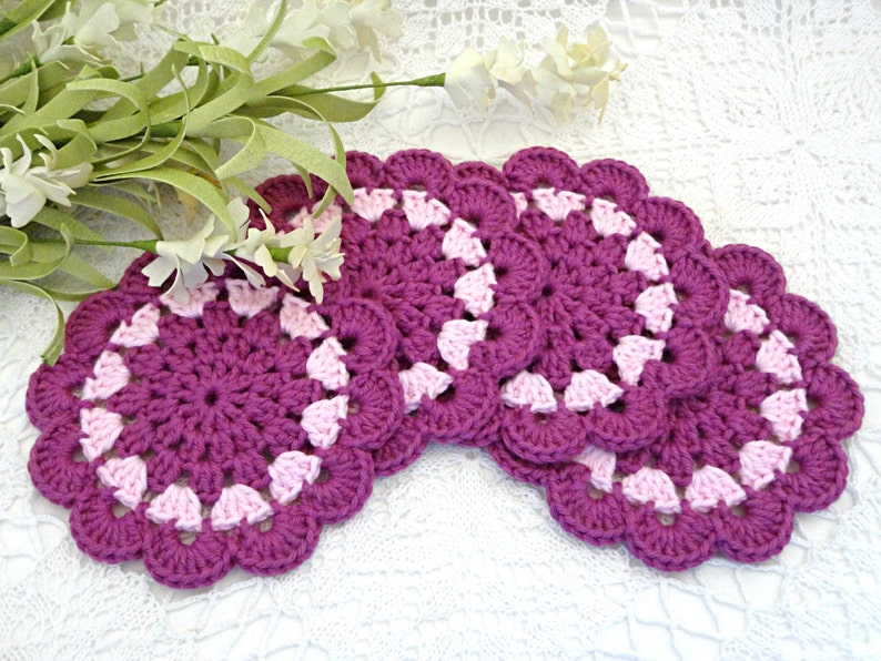May include: Set of five crocheted coasters in shades of pink and purple. The coasters have a scalloped edge and a center design of pink and purple circles.