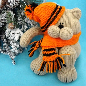 Peut inclure: Un ours en peluche beige tricot&eacute; portant un bonnet et une &eacute;charpe ray&eacute;s orange et noir. L'ours est assis sur un fond bleu.