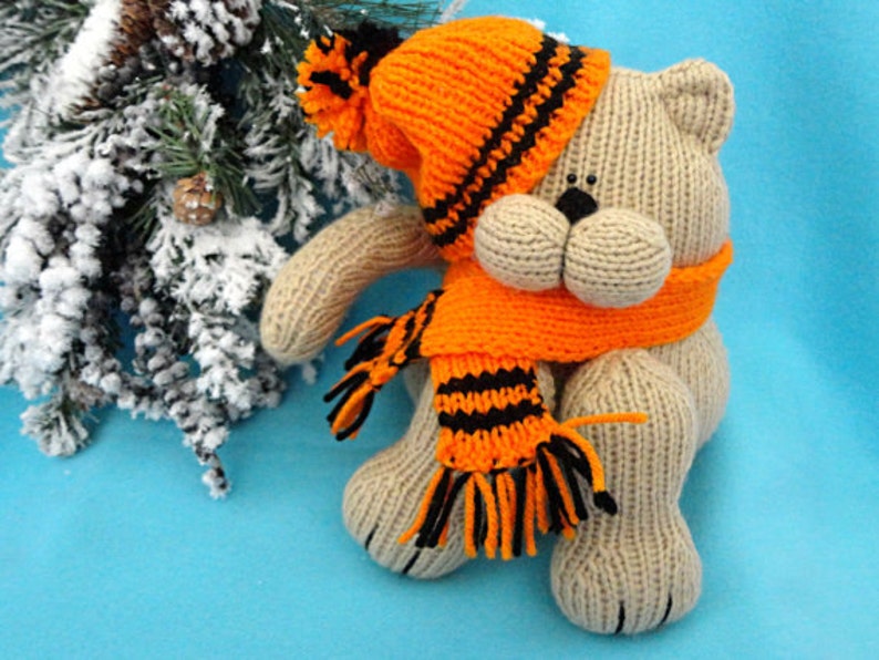 Peut inclure: Un ours en peluche beige tricot&eacute; portant un bonnet et une &eacute;charpe ray&eacute;s orange et noir. L'ours est assis sur un fond bleu.