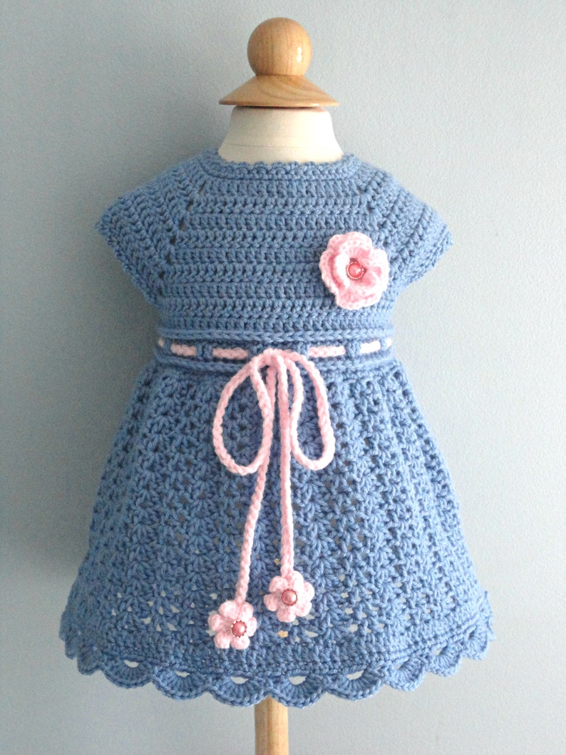 May include: A blue crocheted baby dress with pink flower embellishments and a pink ribbon tie.