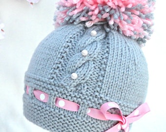 May include: Knitting pattern PDF for a gray knitted hat with a pink and gray pom pom, white pearl embellishments, and a pink ribbon with white polka dots.