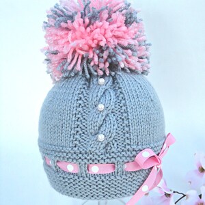 Puede incluir: Sombrero de beb&eacute; de punto gris con un pomp&oacute;n rosa y gris en la parte superior. El sombrero tiene un patr&oacute;n de punto de cable y est&aacute; decorado con botones blancos perlados. Una cinta rosa con lunares blancos est&aacute; atada alrededor del sombrero.