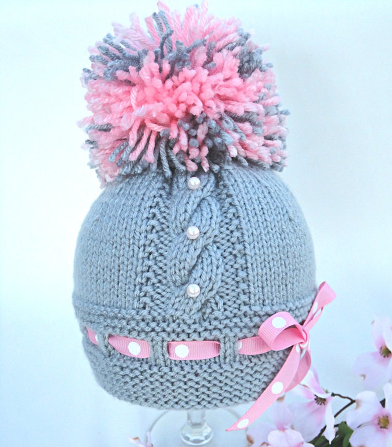 Puede incluir: Sombrero de beb&eacute; de punto gris con un pomp&oacute;n rosa y gris en la parte superior. El sombrero tiene un patr&oacute;n de punto de cable y est&aacute; decorado con botones blancos perlados. Una cinta rosa con lunares blancos est&aacute; atada alrededor del sombrero.