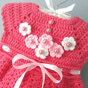 May include: A pink crocheted baby dress with white and pink crocheted flowers and pink pearl-like buttons.