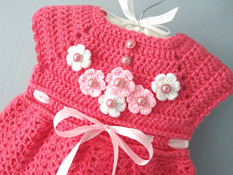 May include: A pink crocheted baby dress with white and pink crocheted flowers and pink pearl-like buttons.