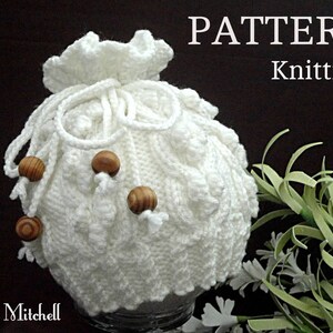 May include: White crocheted drawstring bag with wooden beads. The bag is knitted in a textured pattern and has a ruffled top. The bag is sitting on a dark surface with green foliage in the background. The text "PATTERN Knitting" is visible in the top right corner of the image.