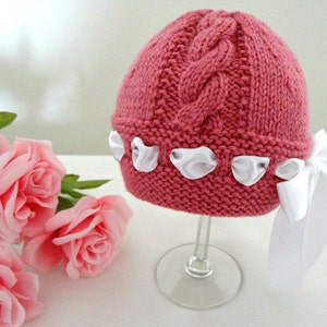 Puede incluir: Un gorro de beb&eacute; de punto rosa con un patr&oacute;n de trenza y lazos de sat&eacute;n blanco.
