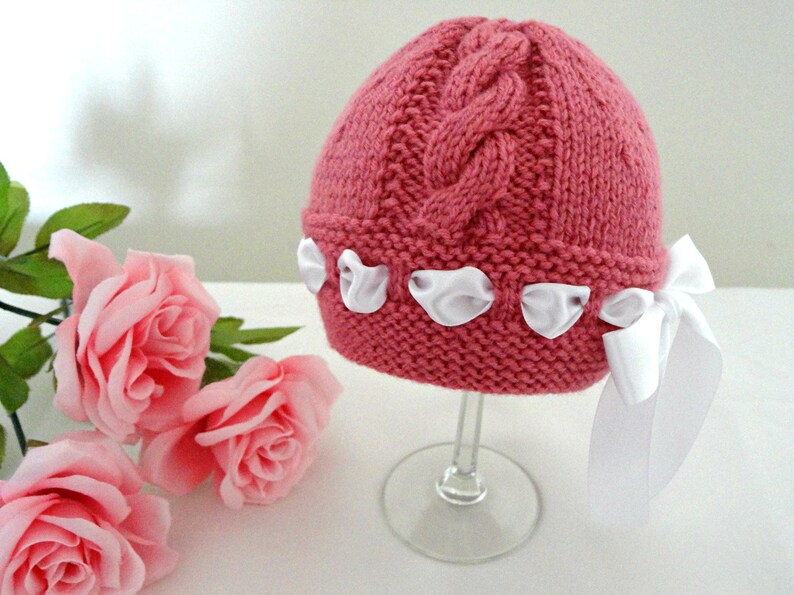 Puede incluir: Un gorro de beb&eacute; de punto rosa con un patr&oacute;n de trenza y lazos de sat&eacute;n blanco.