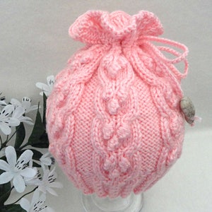 May include: A pink knitted drawstring bag with a cable knit pattern and a small white button.