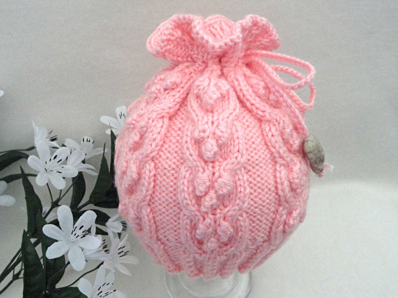 May include: A pink knitted drawstring bag with a cable knit pattern and a small white button.