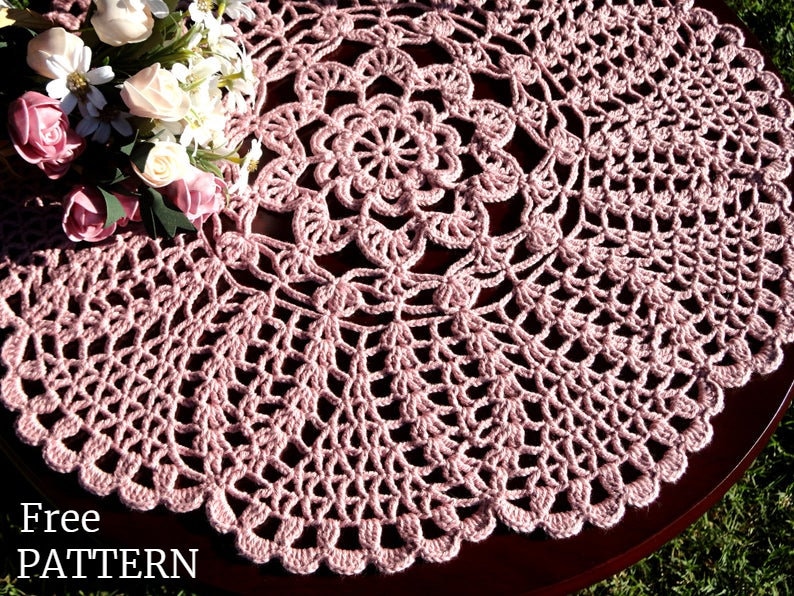 Peut inclure: Un napperon en crochet rose avec un motif floral d&eacute;licat. Le napperon est pos&eacute; sur une table en bois avec un bouquet de fleurs blanches et roses en arri&egrave;re-plan. Mod&egrave;le gratuit.