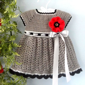 May include: A grey crocheted baby dress with a red poppy flower and a silver ribbon. The dress has a black and white trim.