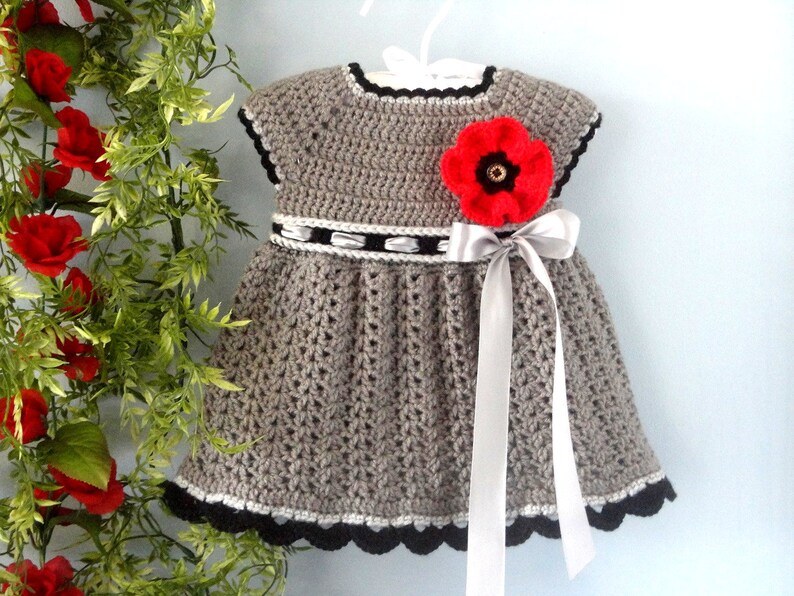 May include: A grey crocheted baby dress with a red poppy flower and a silver ribbon. The dress has a black and white trim.