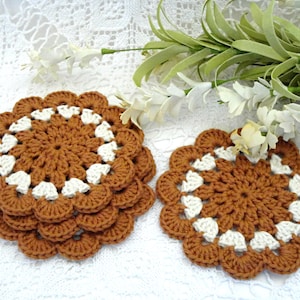 May include: Set of four crocheted coasters in brown and white. The coasters are round and have a scalloped edge. They are made with a delicate crochet pattern.