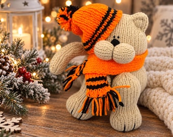 Op de afbeelding: Een beige gebreid knuffelkatje met een textuur, voorzien van een feloranje en zwart gestreepte gebreide muts met een bijpassende pompon en een bijpassende oranje en zwart gestreepte sjaal met franjes aan de uiteinden, zittend rechtop.