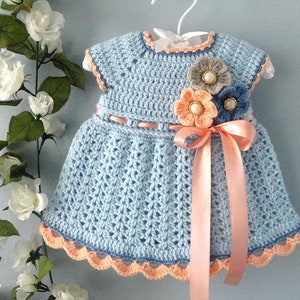 May include: A light blue crocheted baby dress with a peach coloured ribbon and three crocheted flowers in peach, grey, and blue.