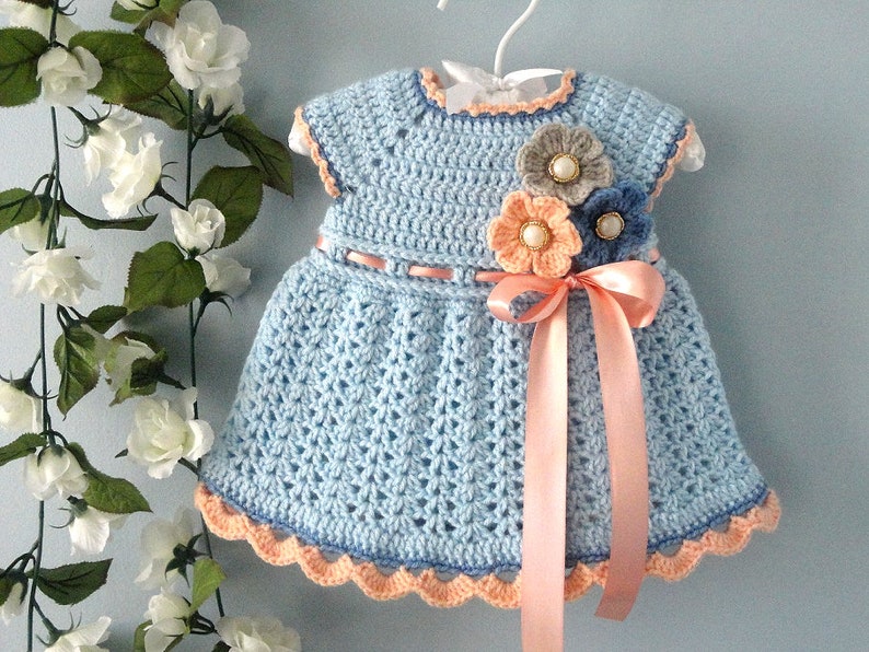 May include: A light blue crocheted baby dress with a peach coloured ribbon and three crocheted flowers in peach, grey, and blue.