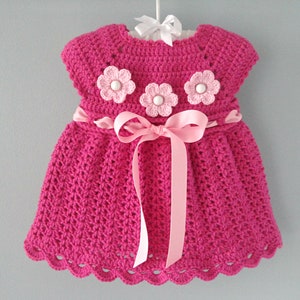 May include: A pink crocheted baby dress with a pink ribbon sash and three pink and white crocheted flowers.