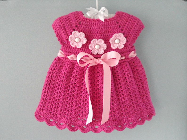 May include: A pink crocheted baby dress with a pink ribbon sash and three pink and white crocheted flowers.