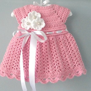 May include: A pink crocheted baby dress with a white crocheted flower embellishment and a white satin ribbon.