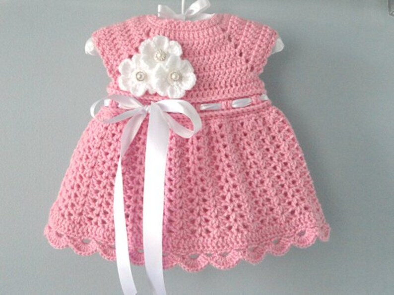 May include: A pink crocheted baby dress with a white crocheted flower embellishment and a white satin ribbon.