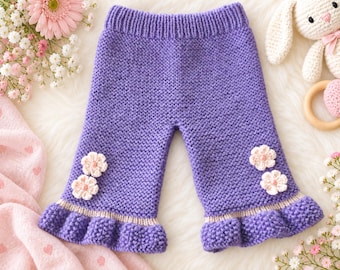 Op de afbeelding: Paarse gebreide broek met een geribbelde tailleband, een ribbelsteek textuur en een geplooide zoom met een lichtbeige accentstreep. Elke broekspijp is versierd met twee kleine, lichtroze gehaakte bloemen op de voorzijde.