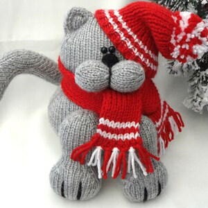 Peut inclure: Un chat en tricot gris portant un bonnet rouge en tricot avec des rayures blanches et une &eacute;charpe rouge en tricot avec des rayures blanches et des glands.