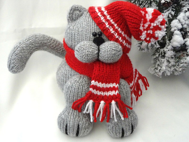 Peut inclure: Un chat en tricot gris portant un bonnet rouge en tricot avec des rayures blanches et une &eacute;charpe rouge en tricot avec des rayures blanches et des glands.