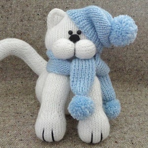 Peut inclure: Jouet en forme de chat blanc tricot&eacute; portant un bonnet et une &eacute;charpe bleus avec des pompons.