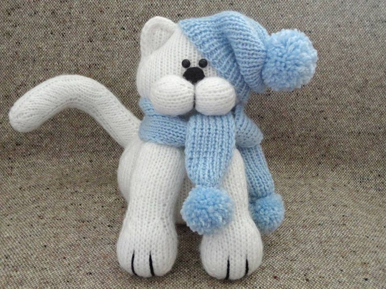 Peut inclure: Jouet en forme de chat blanc tricot&eacute; portant un bonnet et une &eacute;charpe bleus avec des pompons.