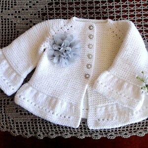 May include: White knitted baby cardigan with a ruffled hem and cuffs. The cardigan has a button closure and a grey flower embellishment.