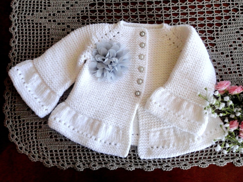 May include: White knitted baby cardigan with a ruffled hem and cuffs. The cardigan has a button closure and a grey flower embellishment.