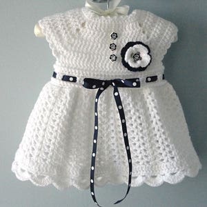 May include: White crocheted baby dress with a black and white polka dot ribbon and a black and white flower embellishment.