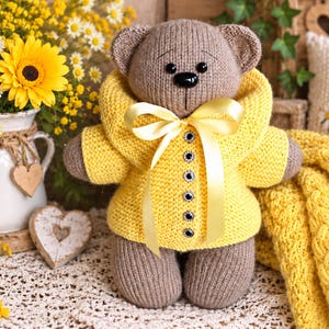 Op de afbeelding: Een lichtbruine gebreide teddybeer met een felgeel vest met een grote kraag, vijf donkere knopen en een gele satijnen strik om de nek. De beer heeft zwarte kraaloogjes en een zwarte neus, afgewerkt met zacht en getextureerd garen.