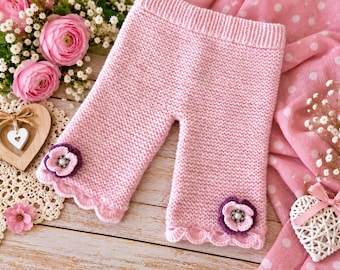 Op de afbeelding: Lichtroze gebreide broek met een geribbelde tailleband en een geschulpte zoom. Elke broekspijp is versierd met een gehaakte bloemapplicatie in paarse en roze tinten, in het midden voorzien van een kleine witte parelachtige kraal.