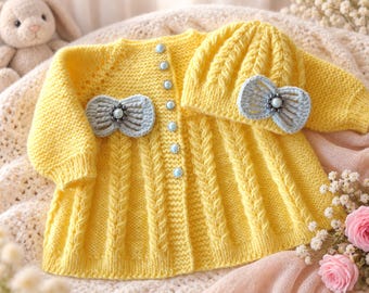 May include: A bright yellow knitted cardigan and matching hat set. The cardigan features vertical cable knit patterns, long sleeves, and a row of light blue buttons. Both the cardigan and the hat are decorated with a light blue crocheted bow.