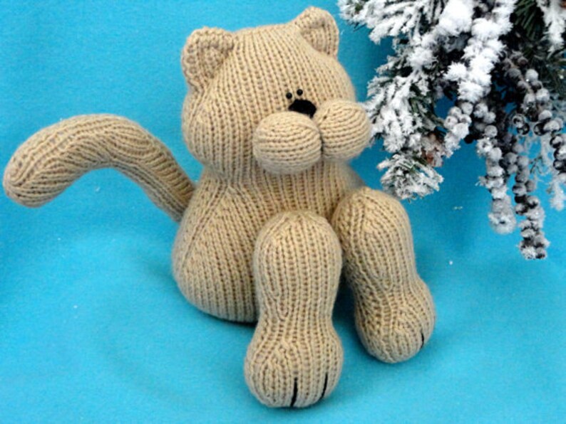 Peut inclure: Un jouet en forme de chat en tricot beige avec des yeux noirs et une queue enroul&eacute;e. Le chat est assis sur une surface bleue.