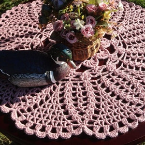 Peut inclure: Un napperon en crochet rose avec un motif d&eacute;coratif, sur lequel est pos&eacute; un canard en bois.