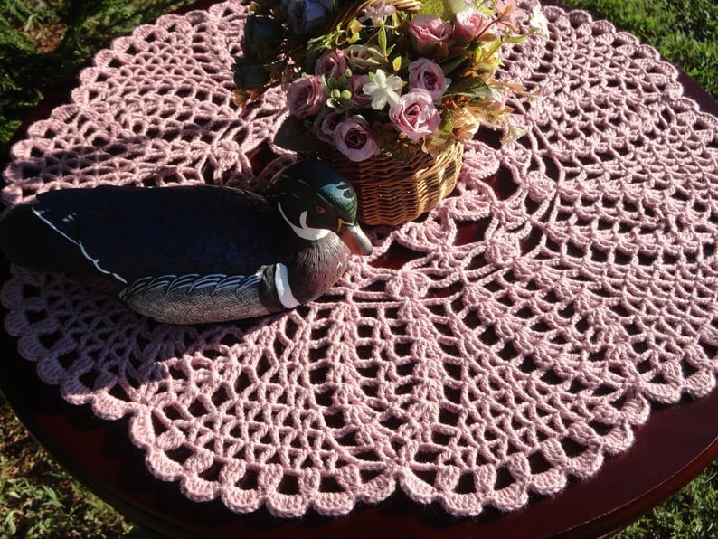 Peut inclure: Un napperon en crochet rose avec un motif d&eacute;coratif, sur lequel est pos&eacute; un canard en bois.