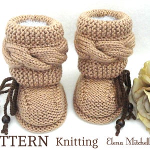 May include: A pair of brown knitted baby booties with a braided cuff and brown leather laces. The booties are sitting on a white surface next to a white rose.