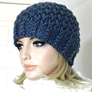 May include: A blue knitted beanie with a textured pattern. The beanie is worn by a person with blonde hair.