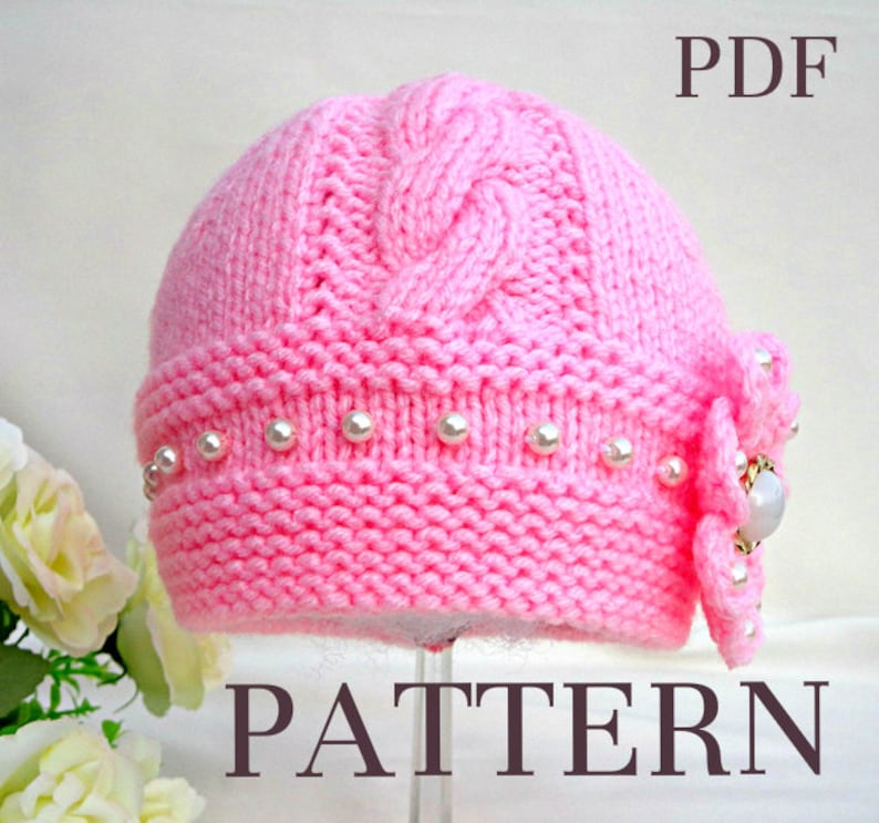 Puede incluir: Un gorro de beb&eacute; de punto rosa con un patr&oacute;n de trenza y un borde de perlas. El gorro tiene un adorno de flor con un centro de pedrer&iacute;a. El texto "PDF PATTERN" est&aacute; impreso debajo del gorro.