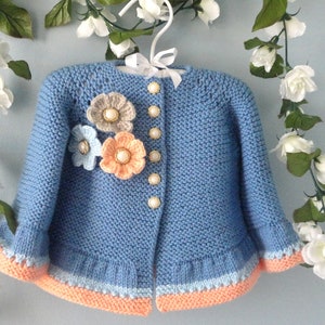 May include: A light blue knitted baby cardigan with a ruffled hem and a floral embellishment on the front. The cardigan has a button closure and is decorated with pearl-like buttons.
