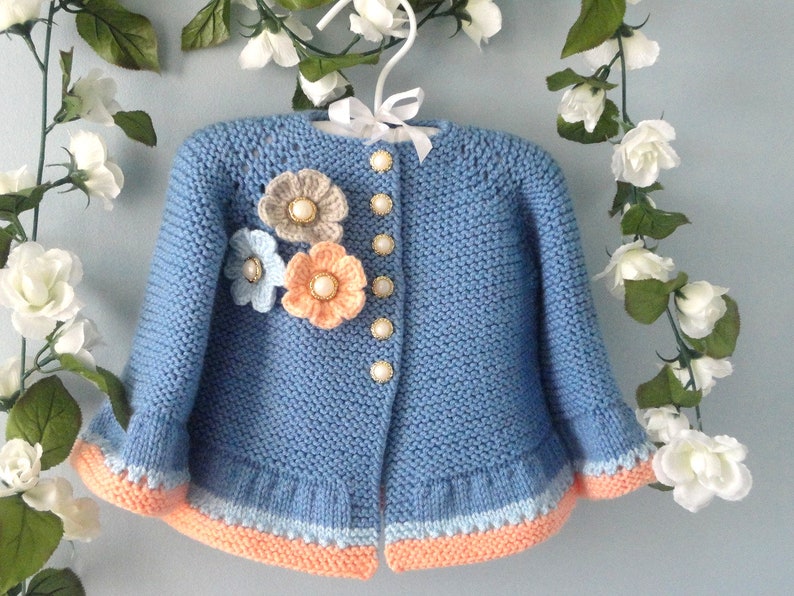 May include: A light blue knitted baby cardigan with a ruffled hem and a floral embellishment on the front. The cardigan has a button closure and is decorated with pearl-like buttons.
