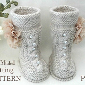 Könnte beinhalten: Ein Paar gestrickte Babyschuhe mit einem Zopfmuster und Perlenbesatz. Jeder Schuh hat eine beige Stoffblume. Der Text "Elena Mitchell Knitting PATTERN PDF" ist im Bild zu sehen.